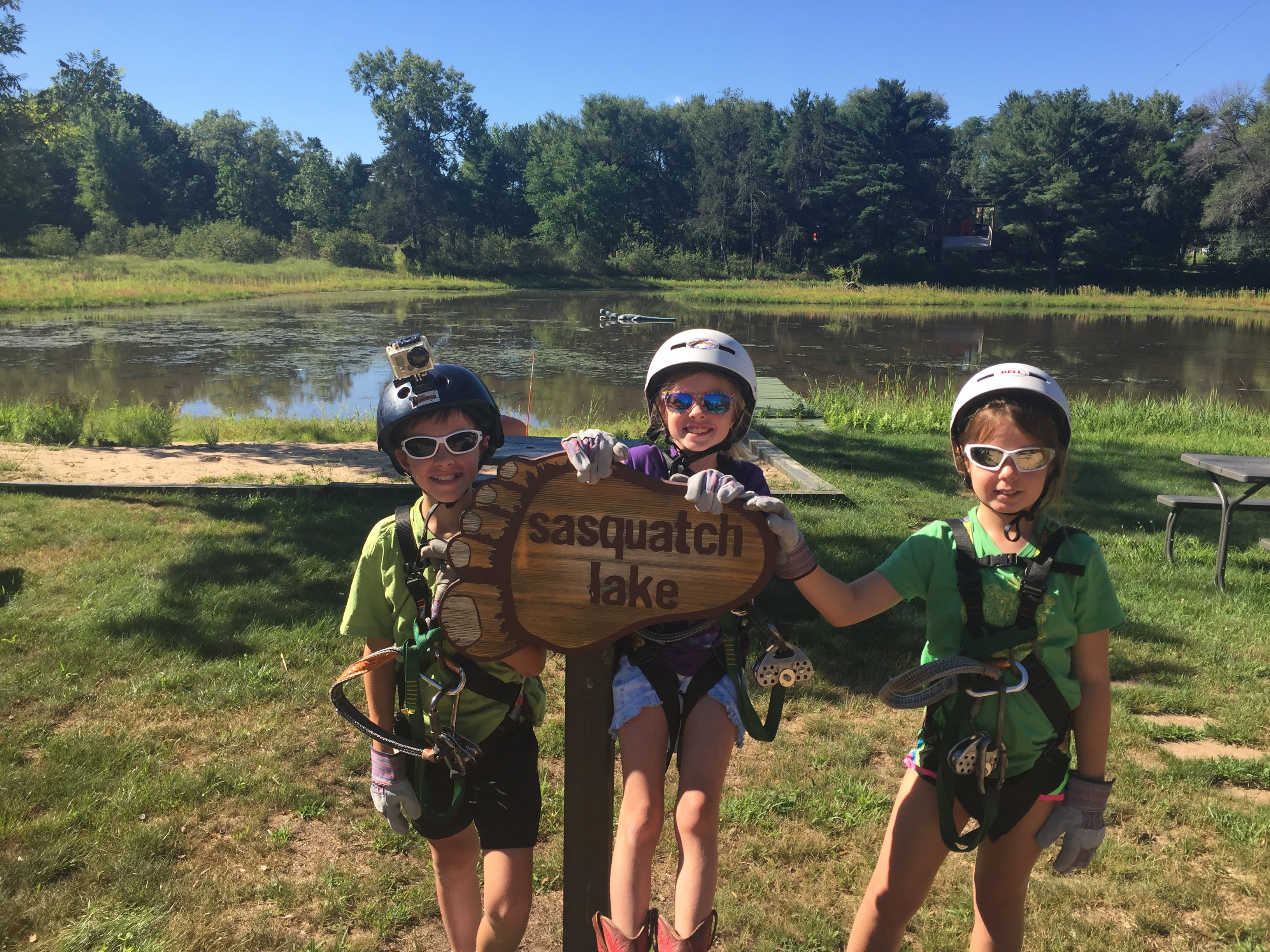 A boy and two girls in zip line gear posing in front of sign for Sasquatch Lake